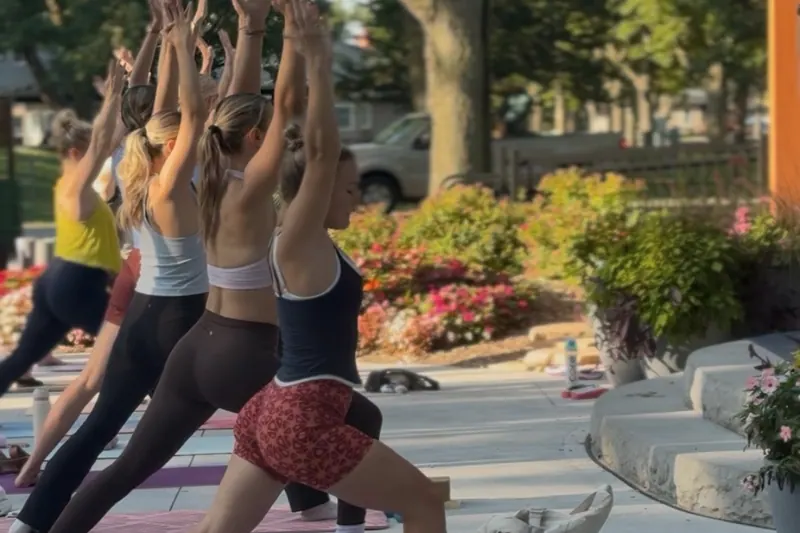 An outdoor power flow class holding warrior two with arms raised overhead beside garden flower beds