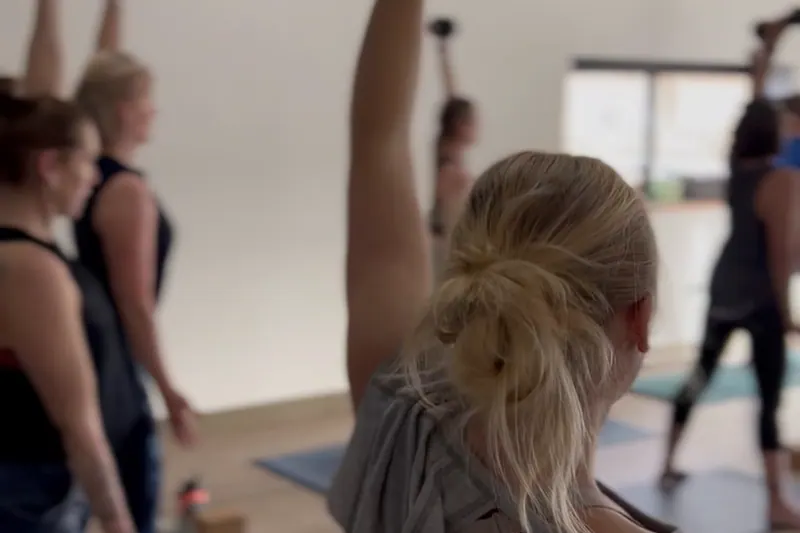 A Flex + Flow class folding into standing side stretches while holding dumbbells overhead, morning light through the studio windows