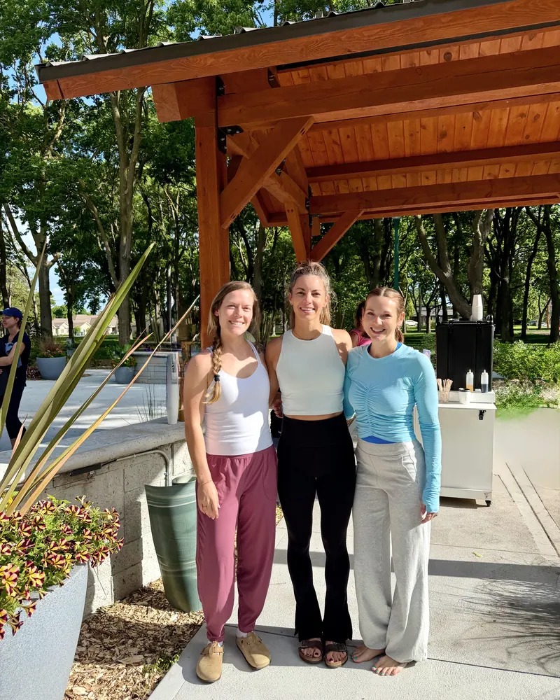 Three women smiling under a timber pavilion after an outdoor class, one barefoot, a coffee cart behind them