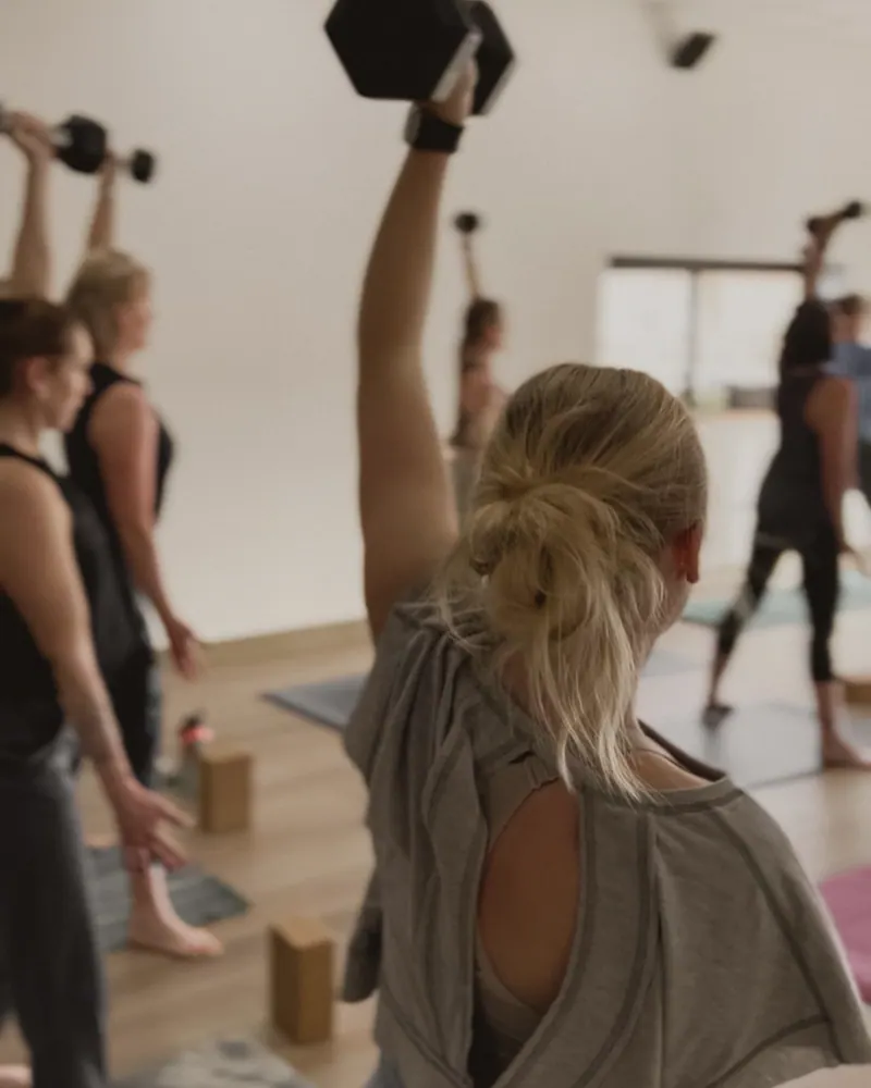 A Built for Her class pressing dumbbells overhead in the studio, seen from behind a member in a grey tee with her hair in a bun, cork blocks on the floor