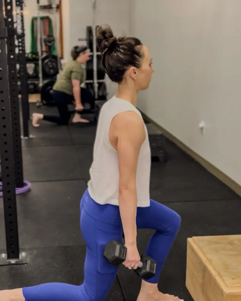 A woman in blue leggings and a white tank holding a dumbbell lunge beside a wooden plyo box, another member kneeling at the squat rack behind her