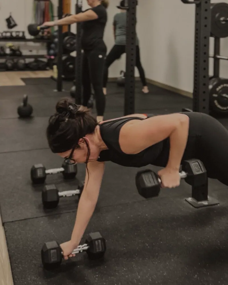A woman in a black tank in a plank on two dumbbells, rowing one to her hip on the black gym floor, two other members training at the rack behind her