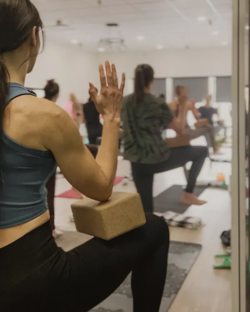 A woman in a teal top in a low lunge with a cork block balanced on her thigh under her elbow, the rest of the class in the same pose in the bright studio behind her
