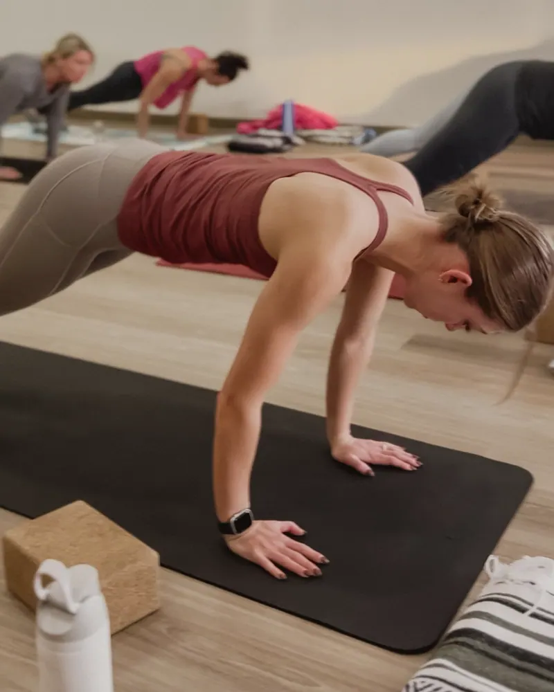 A woman in a maroon tank holding a high plank on a black mat, a cork block and water bottle beside her, three classmates planking behind