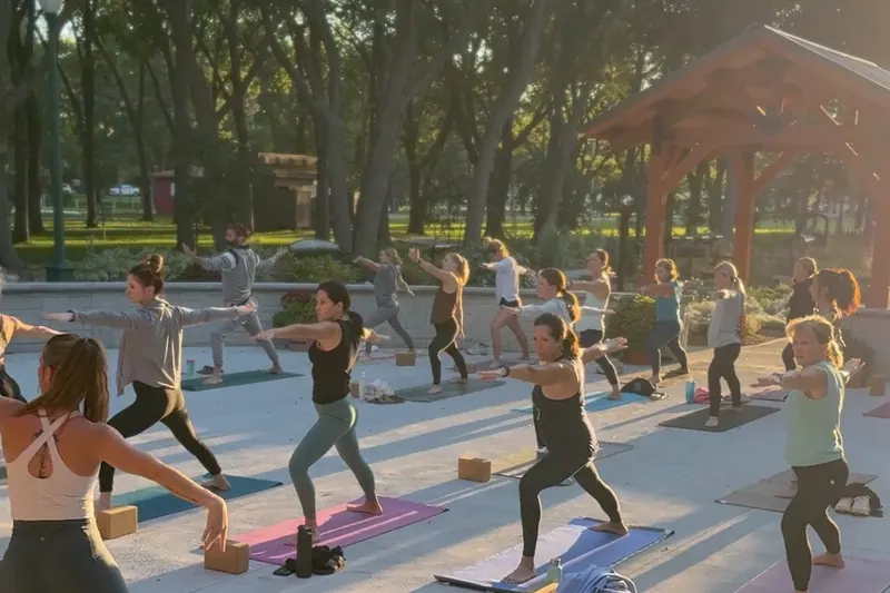 An evening outdoor yoga class reaching upward on mats at a park pavilion, the sun setting through the trees behind them